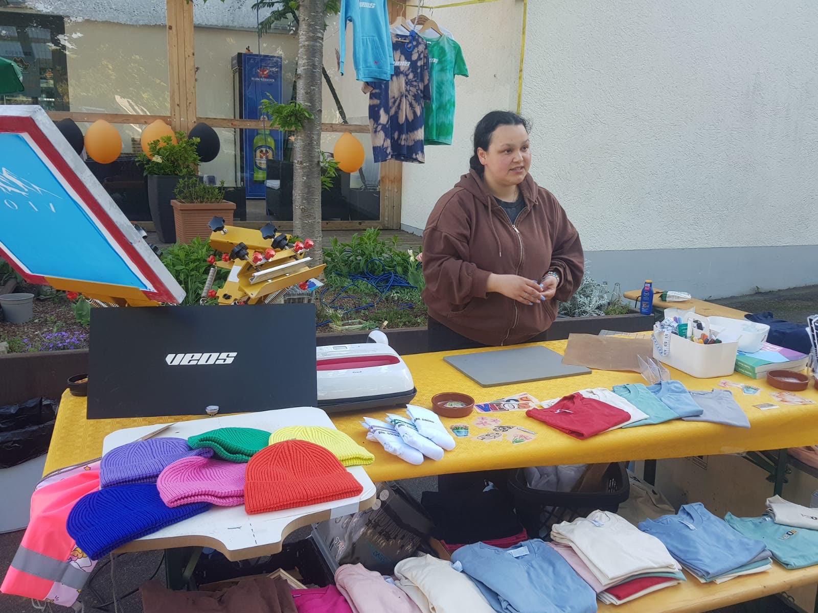 Der Stand von VEOS im Freien: ein gelb gedeckter Tisch mit bunten Mützen, Shirts und einer Druckpresse, dahinter eine Frau.
