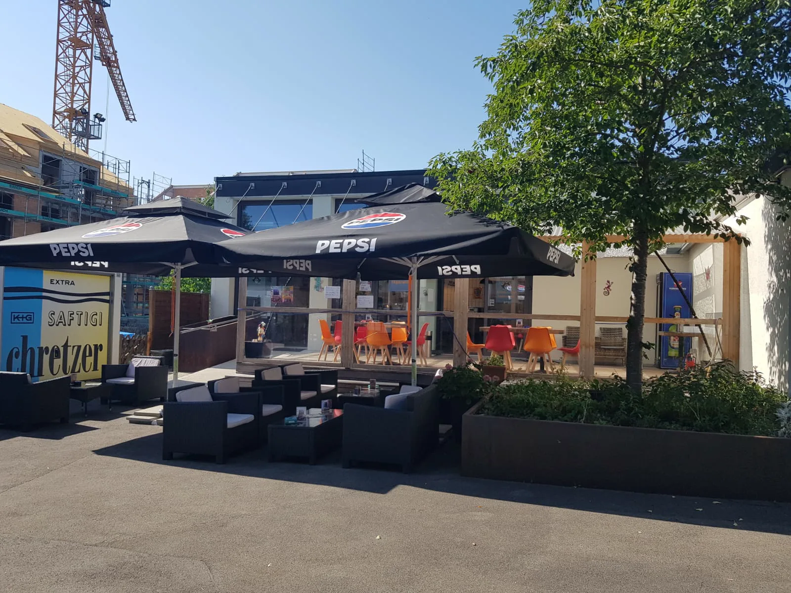 Das Pulfer-fass von aussen an einem sonnigen Tag: zwei große schwarze Sonnenschirme über Loungesesseln auf dem Vorplatz, dahinter die verglaste Front mit orangen und roten Stühlen, rechts ein Baum und ein bepflanzter Trog.