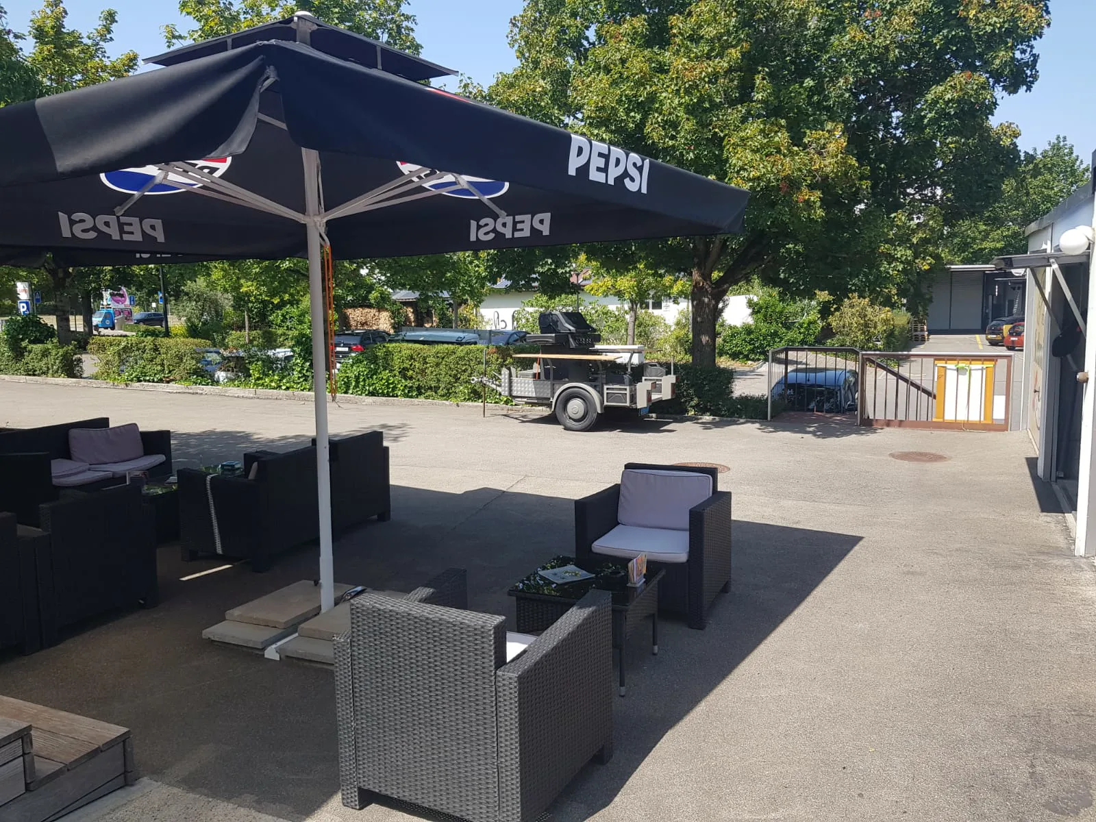 Der Aussensitzplatz mit schwarzen Sonnenschirmen über Lounge-Möbeln, daneben ein Hochbeet mit Bepflanzung.