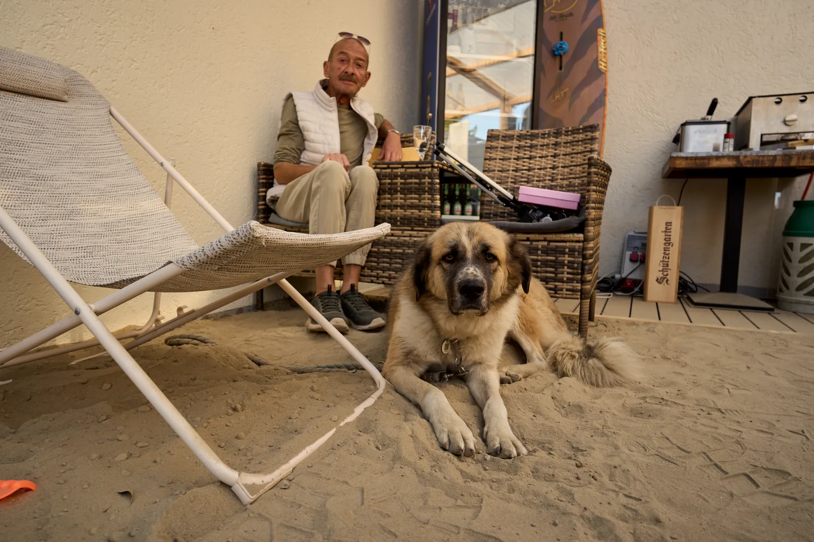 Ein grosser Hund liegt im Sand vor einem Liegestuhl, dahinter sitzt sein Mensch.