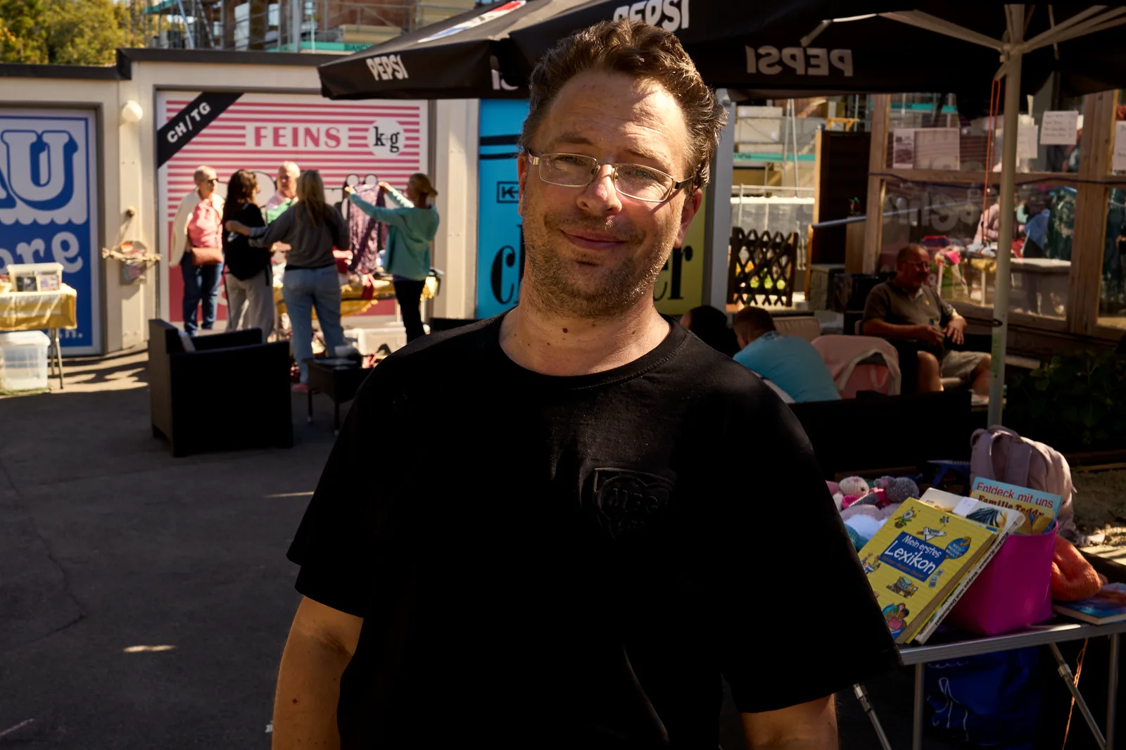 Ronny, der Besitzer des Pulfer-fass, im schwarzen T-Shirt, lächelt in die Kamera, hinter ihm Stände und Schirme.