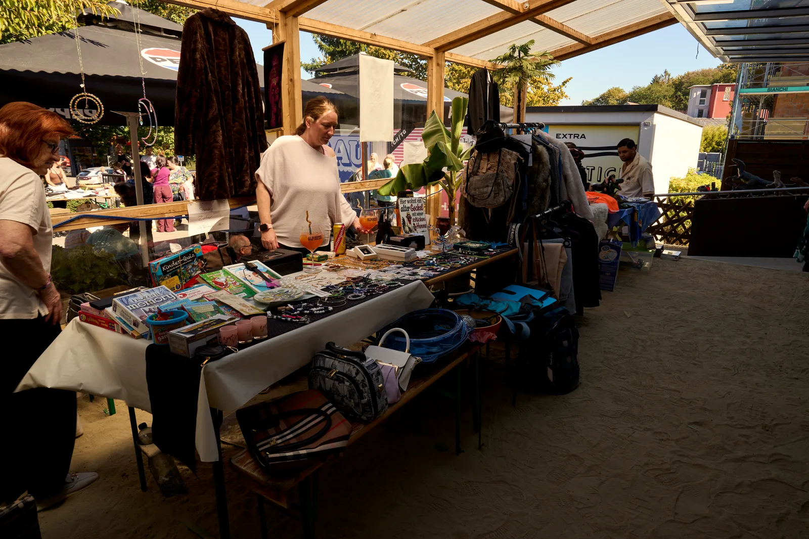 Ein Stand unter dem Holzdach der Terrasse: Tische voller Taschen, Kleider und Krimskrams, zwei Verkäuferinnen.