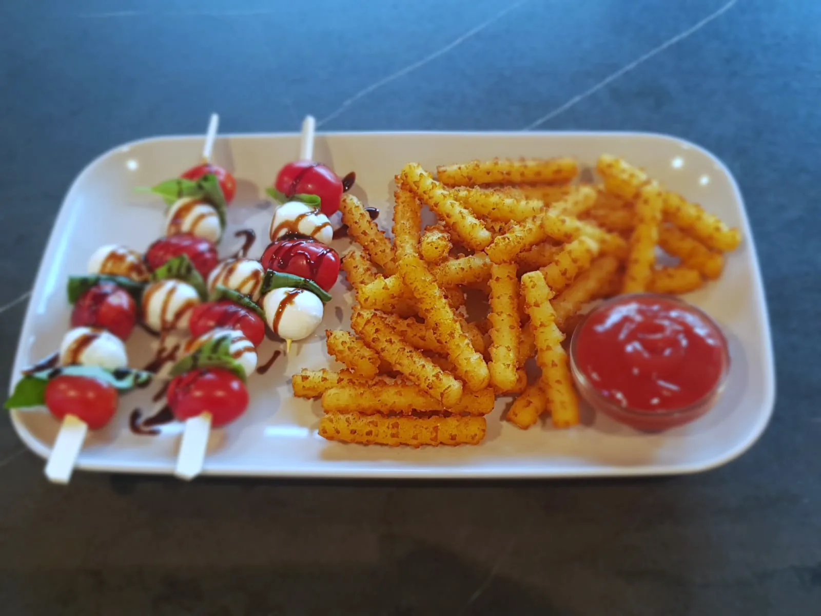 Tomaten-Mozzarella-Spiesse mit Balsamico auf einem weissen Teller, daneben eine Portion Crispy Fries und ein Schälchen Ketchup.