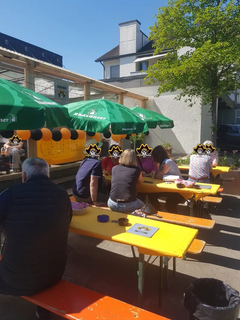 Der Aussensitzplatz an einem Sonnentag: besetzte Biertische mit gelben Decken unter grünen Sonnenschirmen, dahinter die überdachte Terrasse mit oranger Girlande.